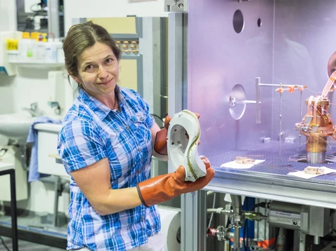 Dr. Eve Stenson hält ein Modell einer nicht-planaren Magnetspule, mit dem supraleitende Bänder getestet werden. Im Hintergrund ist der Prüfstand zu sehen.